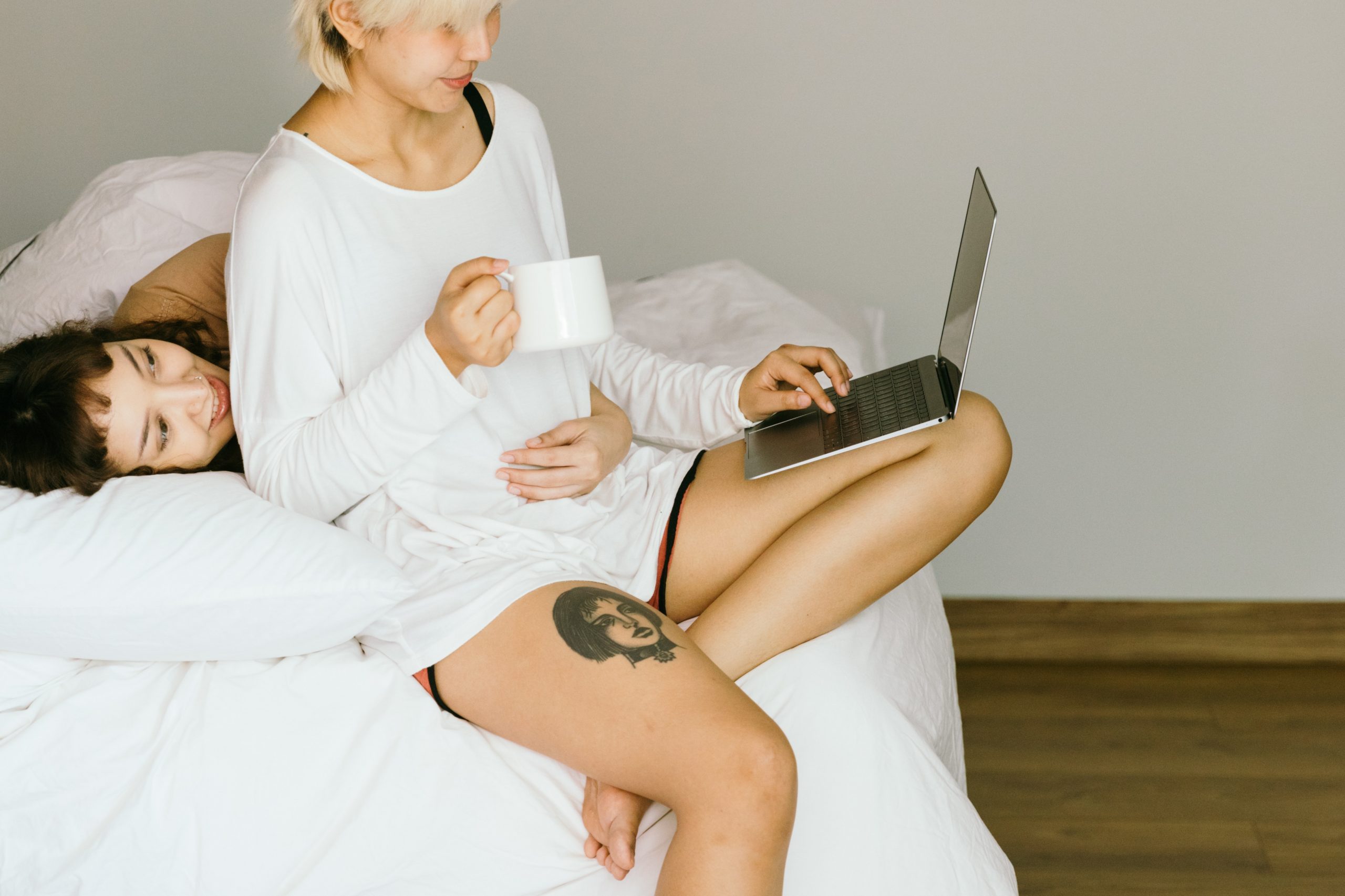 a woman wraps her arm around another woman in bed while she looks at her laptop