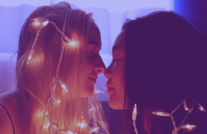 two woman draped in string lights touch noses